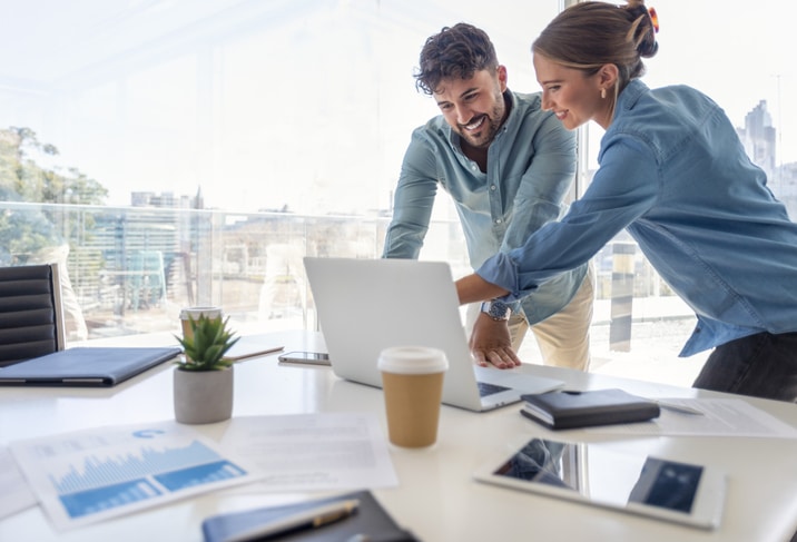 Businessman and businesswoman working on a laptop computer. They are wearing matching casual clothes that could be a uniform and are happy and smiling. They are standing in a board room with a window behind them